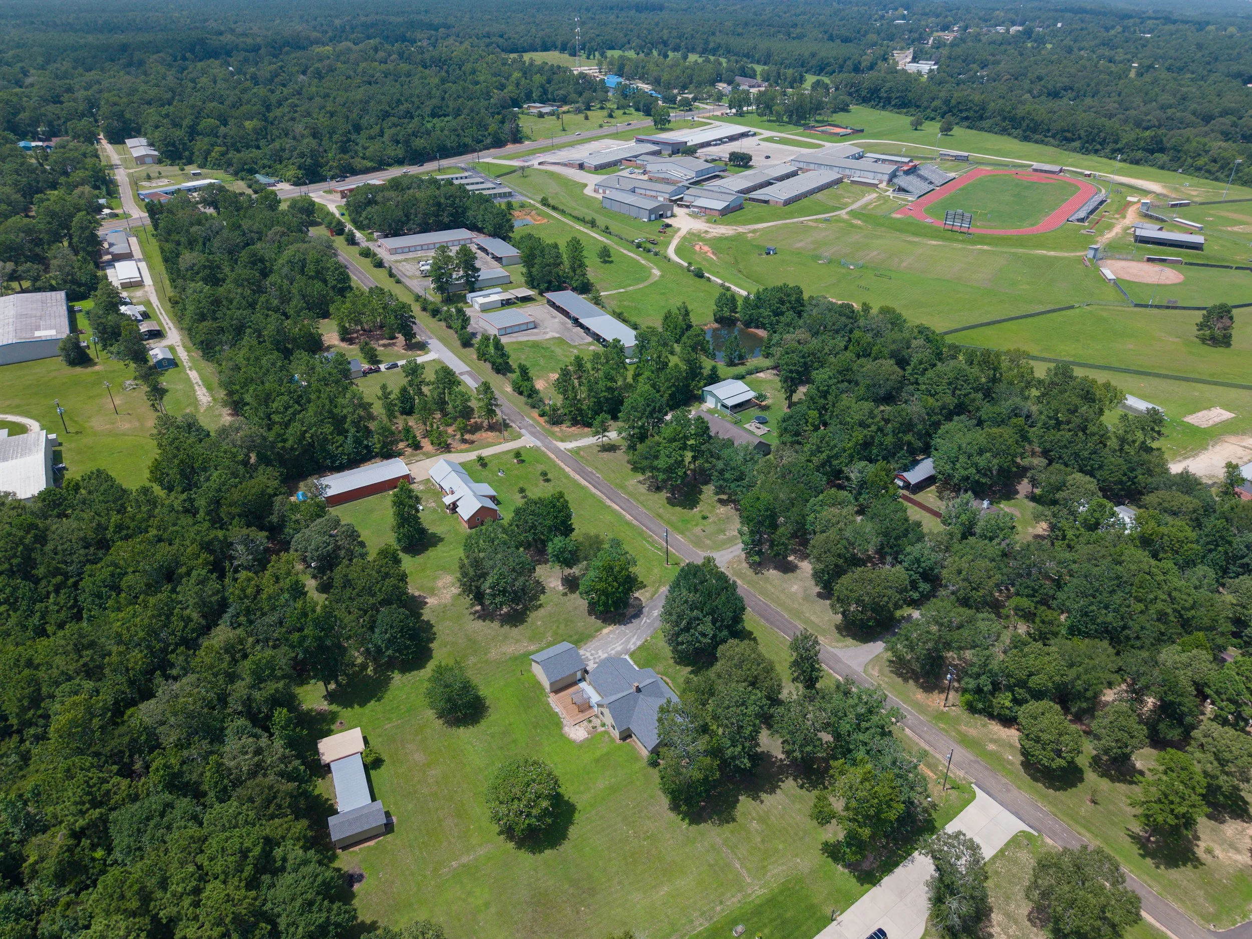 An aerial view of a school campus with multiple buildings, sports fields including a track and baseball field, surrounded by trees and greenery.