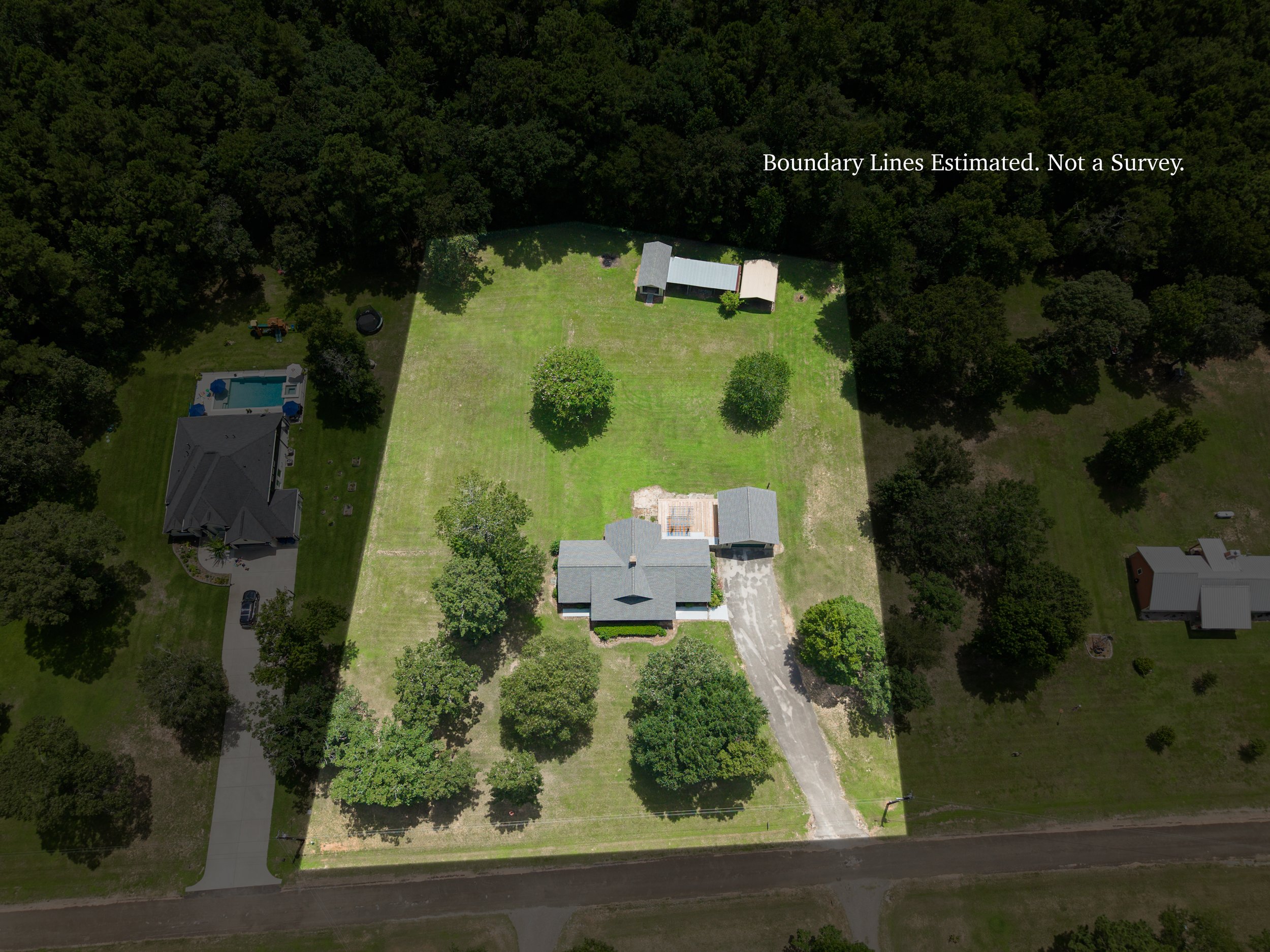 Aerial view of a backyard with a lawn, a small pool, trees, and a wooden shed, surrounded by neighboring houses and dense forest.