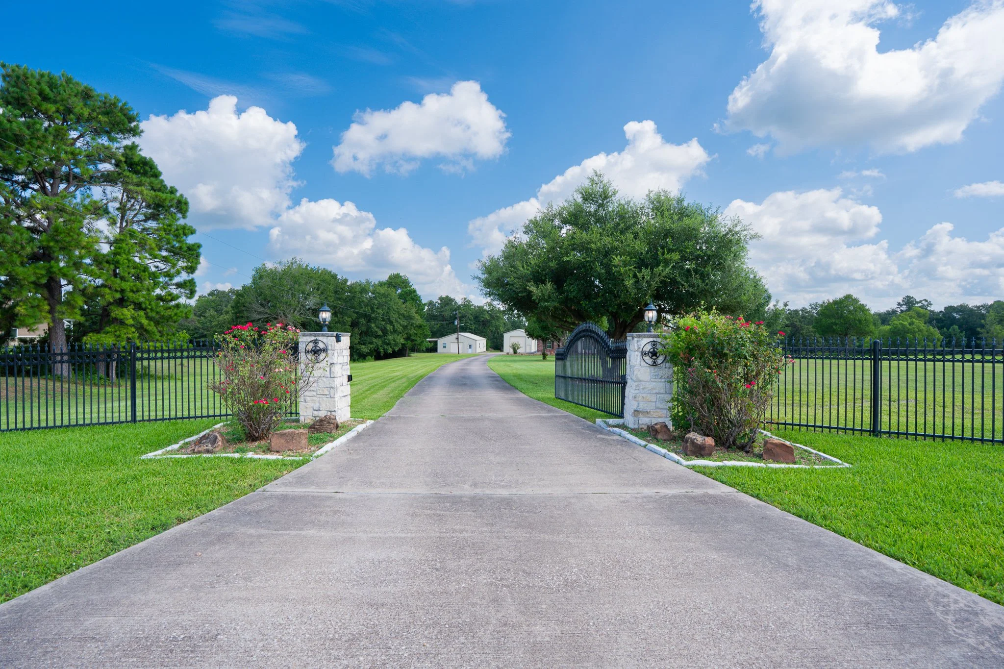 A paved driveway leading to a gate with white stone pillars, surrounded by green grass and colorful flowers, with large trees and a blue sky with scattered clouds in the background.