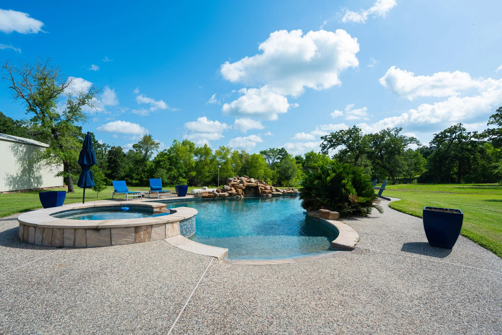 Backyard with a swimming pool, lounge chairs, umbrellas, and a clear blue sky with clouds.