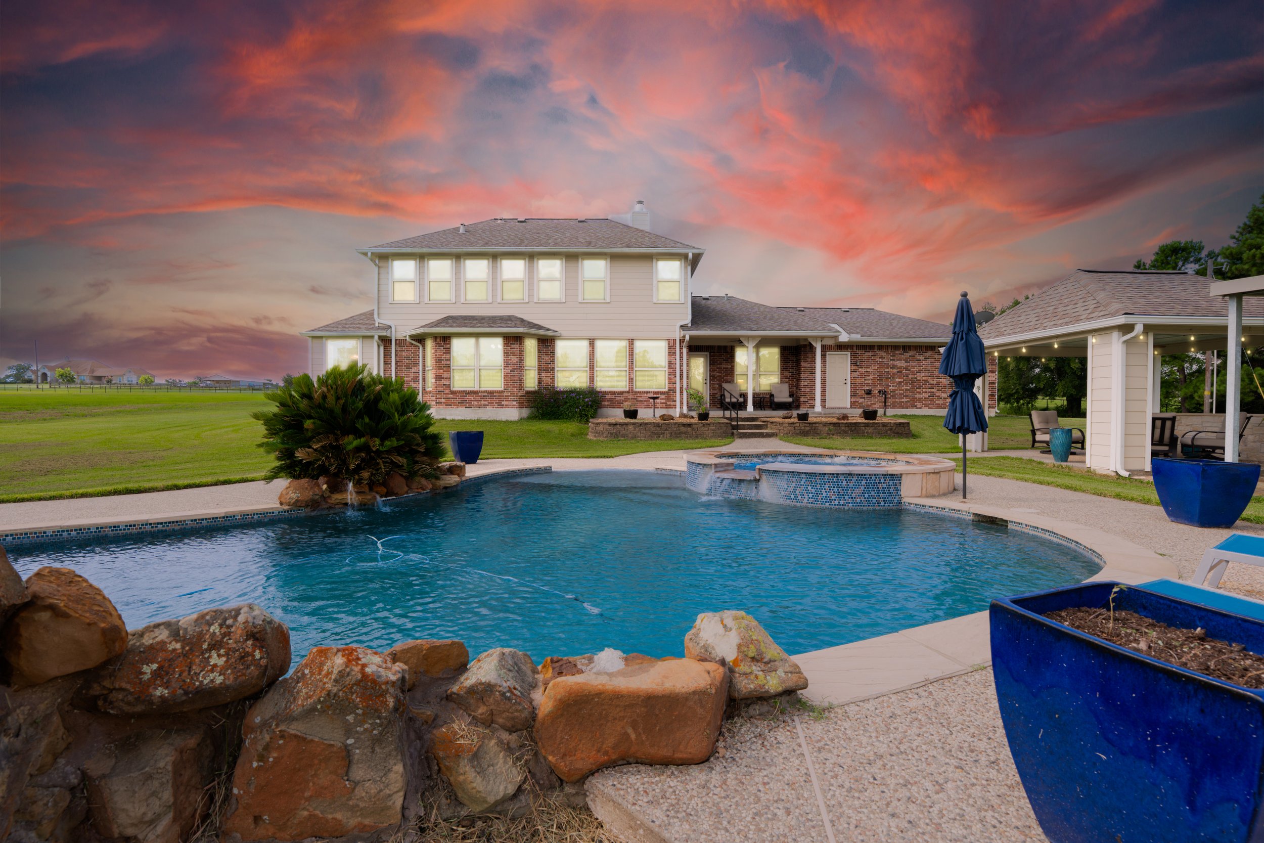 A backyard scene during sunset featuring a swimming pool, patio furniture, a house with large windows, a grassy lawn, and a colorful sky with orange and pink clouds.