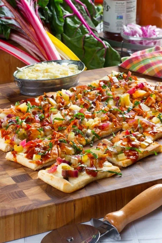 Prepared flatbread pizza topped with assorted vegetables and barbecue sauce on a wooden cutting board. In the background, there are fresh rhubarb, a bowl of shredded cheese, canned ingredients, and a checkered cloth.