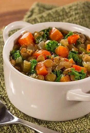 A white bowl filled with vegetable stew containing carrots, green beans, peas, celery, and other mixed vegetables, placed on a green textured cloth with a spoon nearby.