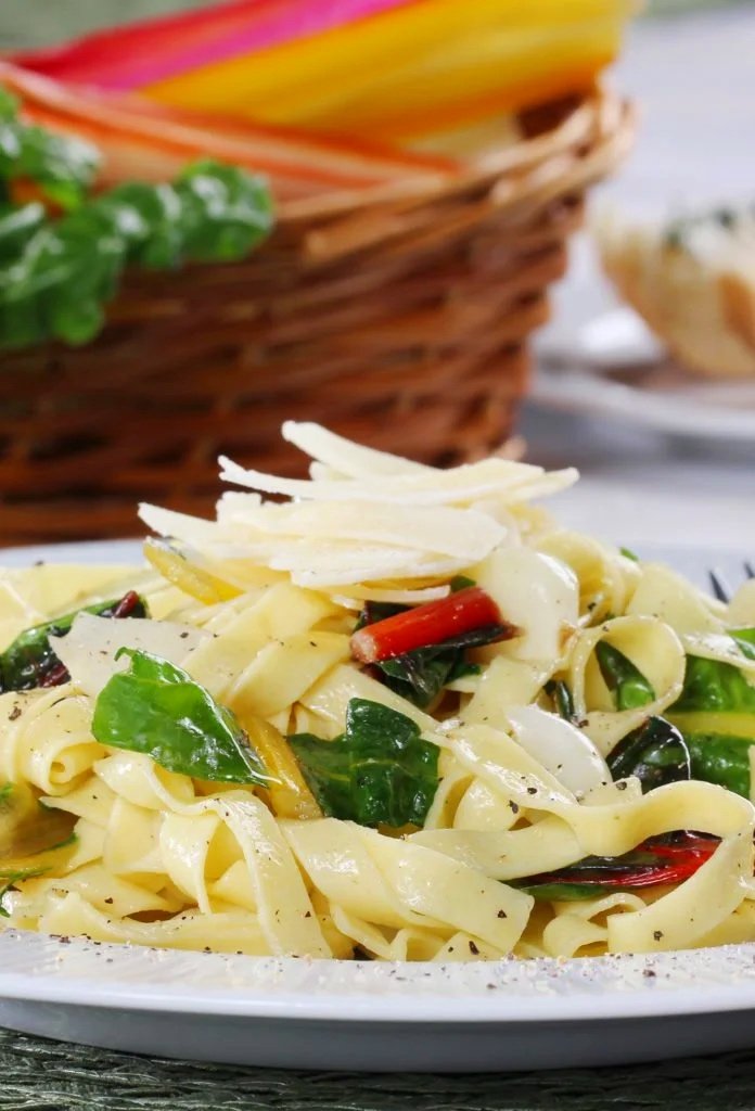 Close-up of a plate of creamy pasta with spinach, red peppers, and grated cheese, with a basket of colorful bread in the background.