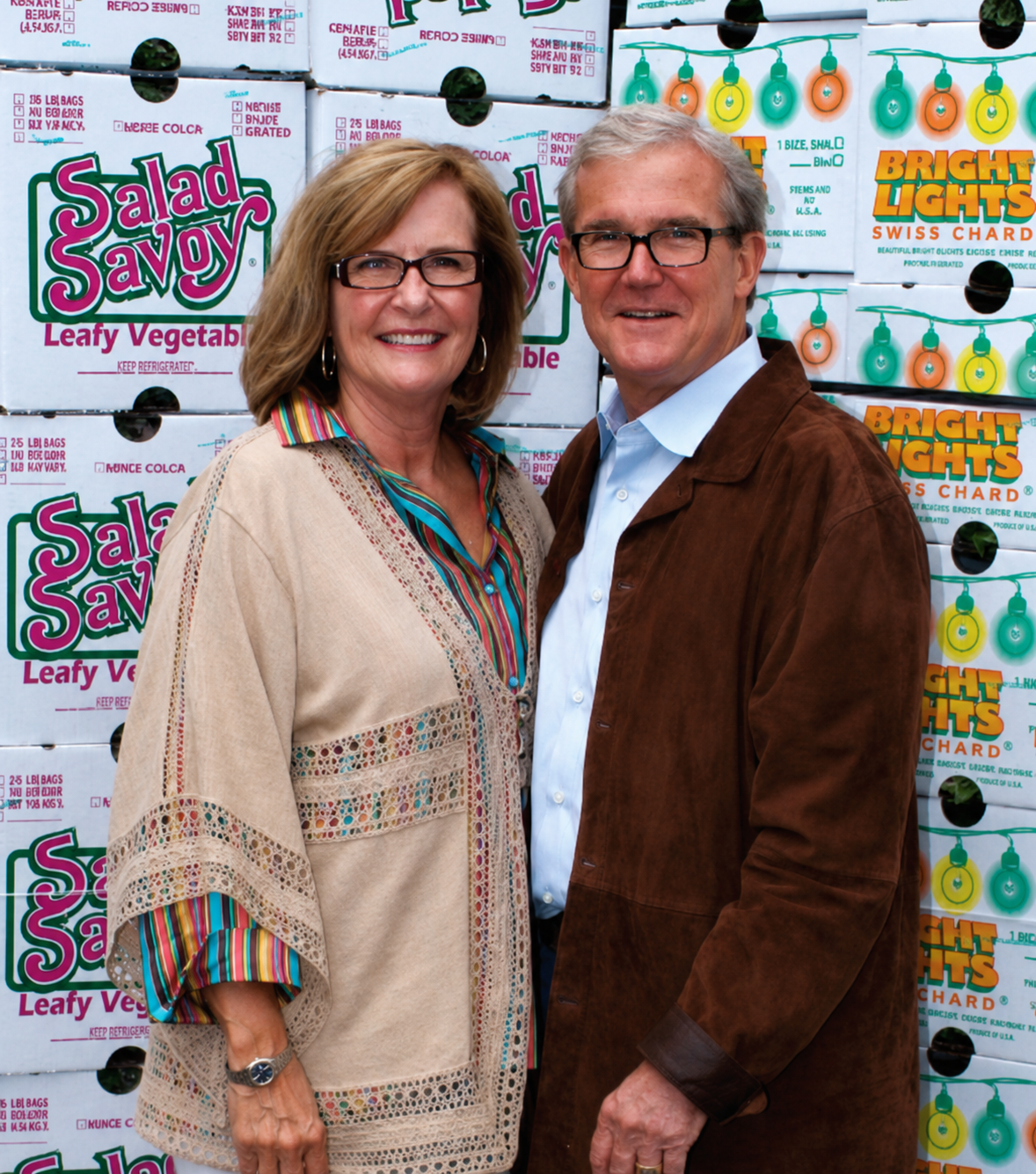 A smiling middle-aged woman and man standing in front of a background of stacked boxes labeled Salad Savoy and Bright Lights Swiss Chard, with colorful light bulb decorations.