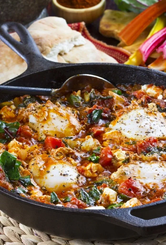 Cast iron skillet with eggs, tomatoes, spinach, and cheese in a sauce.