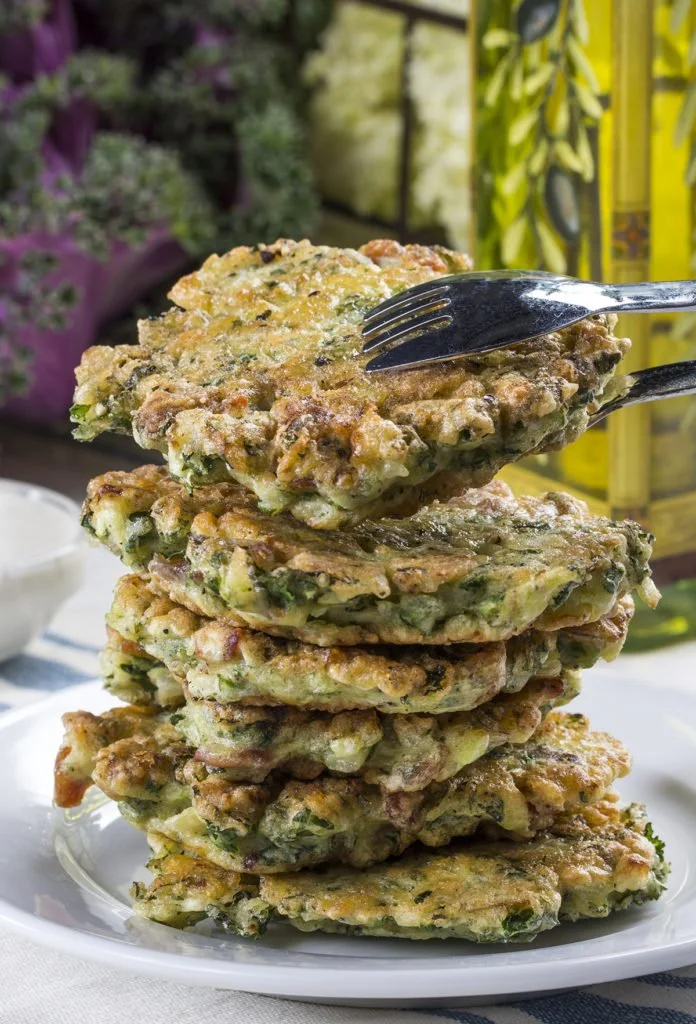A stack of vegetable fritters on a white plate with a fork picking up one fritter, background of blurred plants and yellow decorations.