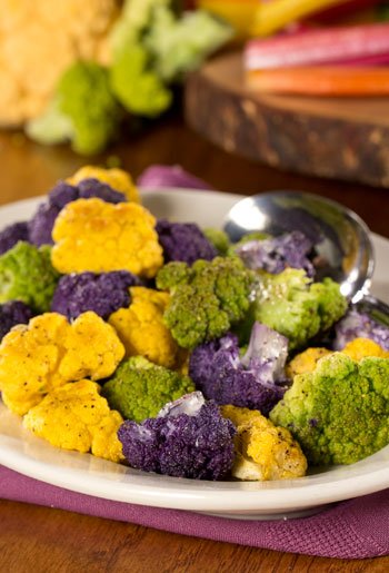 Colorful multicolored cauliflower florets on a white plate with a spoon, set on a purple napkin with a wooden background.