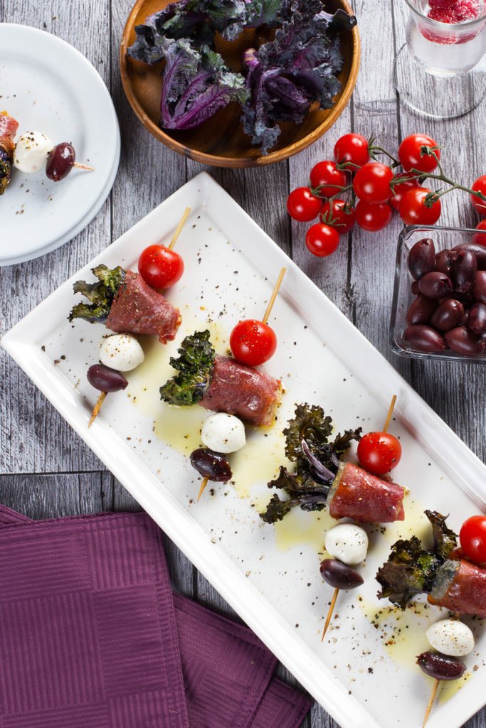 Appetizer plate with prosciutto-wrapped melon balls, cherry tomatoes on skewers, mozzarella balls, and black olives. Surrounding bowls contain mixed greens, grape tomatoes, and black grapes, with a glass of sparkling water garnished with a raspberry.