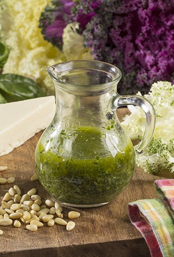 A glass pitcher filled with green salsa on a wooden surface, with fresh flowers and white beans around it.