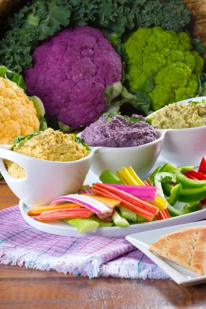 Colorful vegetables, including cauliflower, broccoli, and celery, with bowls of hummus or dips.