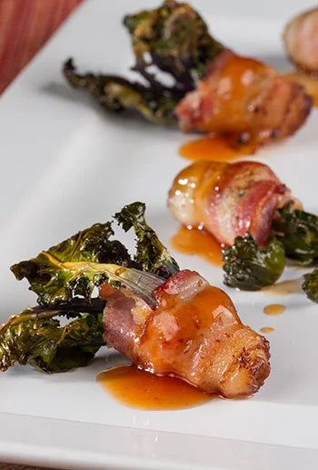 Bacon-wrapped jalapeño poppers with melted cheese and fried kale on a white plate.