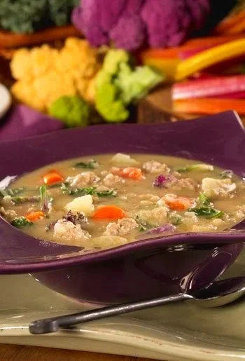 A bowl of vegetable soup with carrots, potatoes, and greens, placed on a table with colorful vegetables in the background.