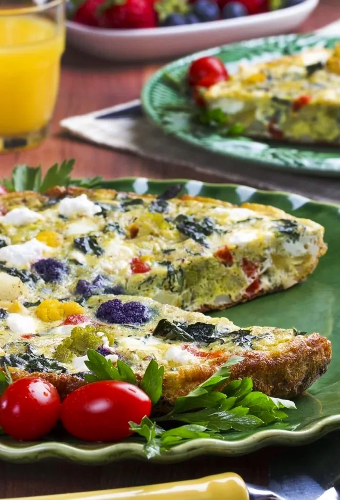 Vegetable frittata on a green plate with cherry tomatoes and parsley, with a glass of orange juice and a fruit salad in the background.