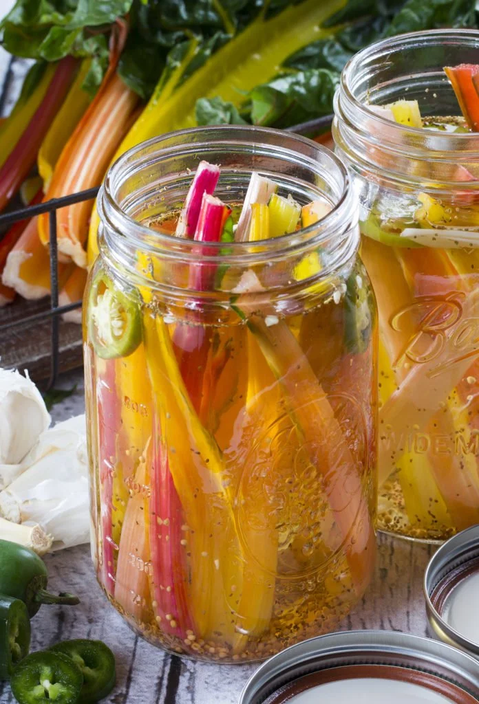 Colorful pickled vegetables in mason jars on a rustic wooden surface, with fresh vegetables and garlic nearby.