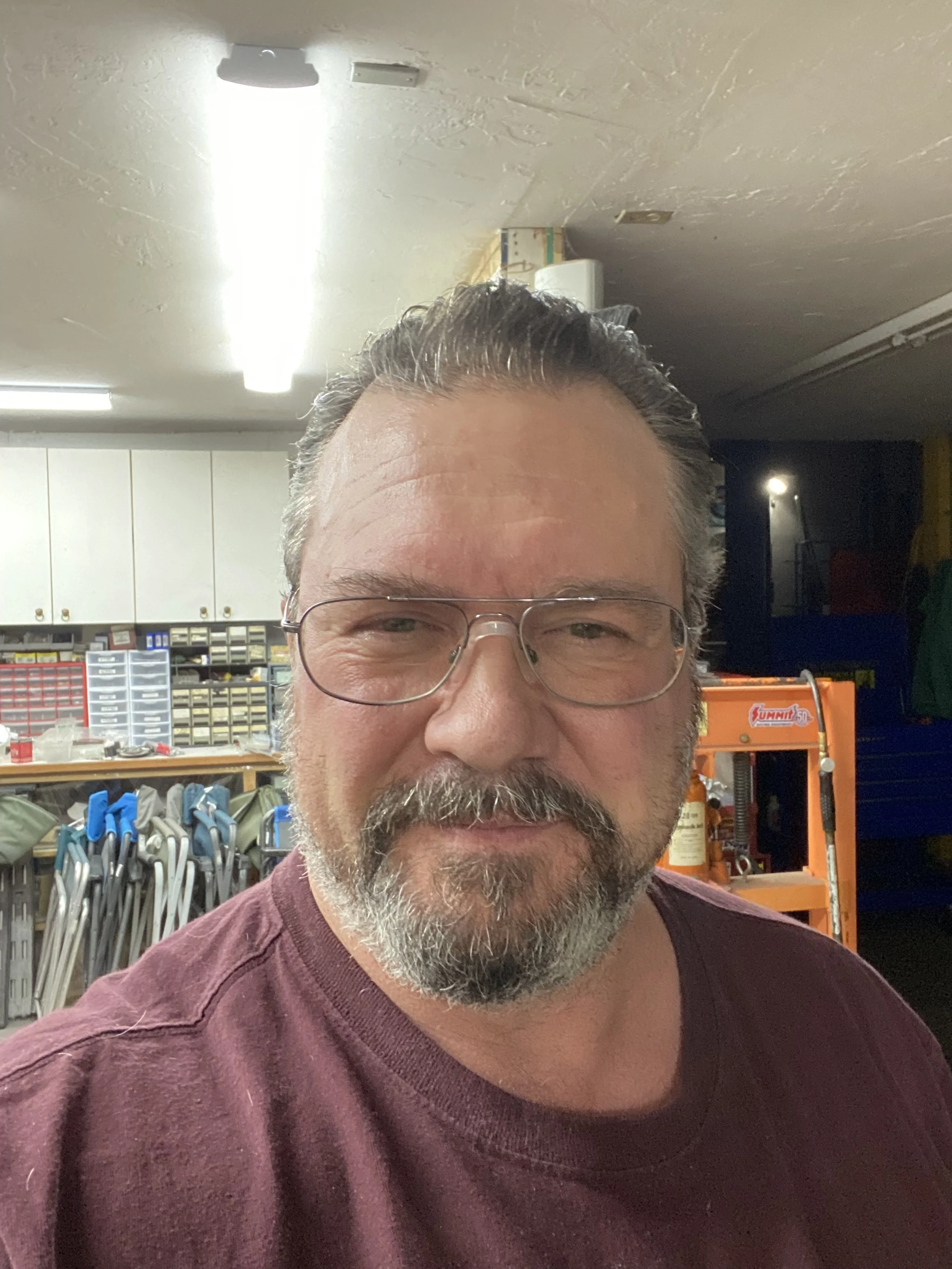 Close-up of a middle-aged man with glasses, gray hair, and a beard, smiling, in a workshop surrounded by tools and equipment.