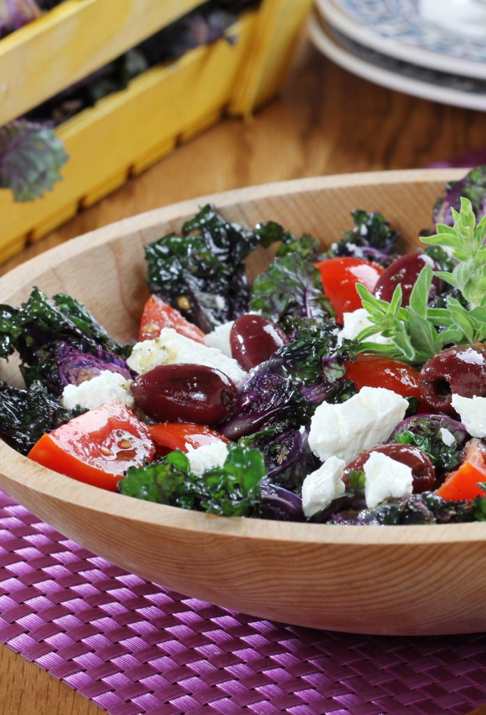 A bowl of salad with kale, cherry tomatoes, red grapes, feta cheese, and fresh herbs on a pink placemat.