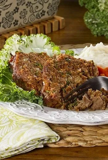 Plate of meatloaf with mashed potatoes and vegetables, garnished with lettuce, on a decorative plate with napkins nearby.