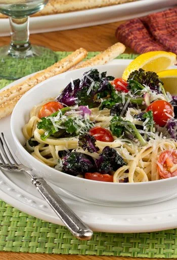 A bowl of pasta salad with cherry tomatoes, mixed greens, grated cheese, and lemon wedges, served with breadsticks on a green placemat.
