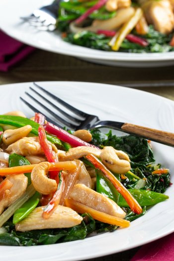 Close-up of a white plate with stir-fried vegetables and chicken, including snow peas, carrots, red peppers, cashews, and greens, with a fork and additional plate in the background.