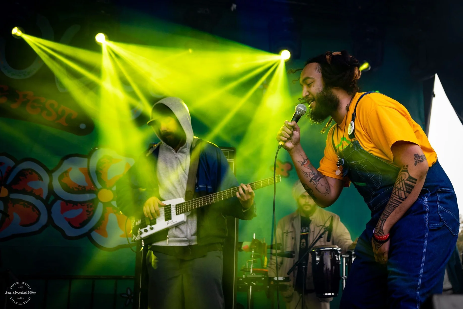 Dread Kennedy performing on stage with yellow lights, one singing into a microphone with tattoos, another playing a keytar, colorful backdrop, and a small audience.