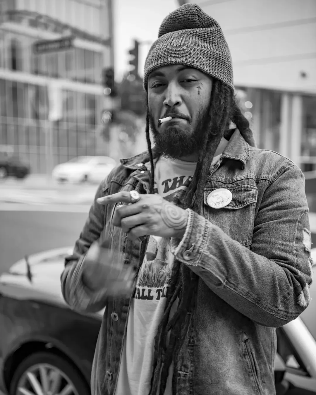 Dread Kennedy in a beanie, denim jacket, and a graphic T-shirt, holding a cigarette in his mouth and pointing with his hand. He is outdoors in an urban setting with blurred buildings and cars in the background.