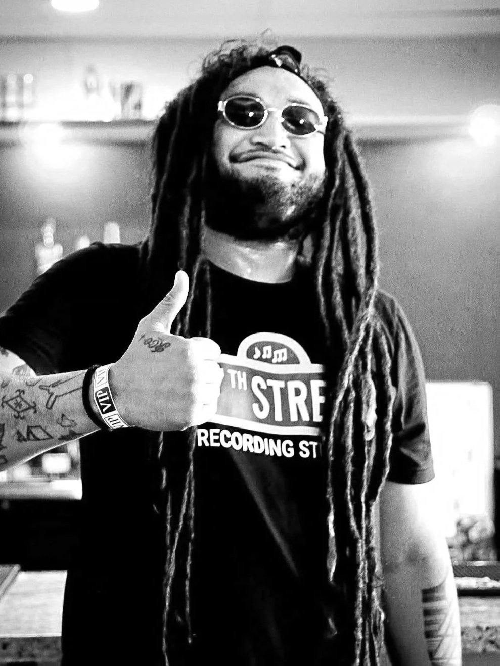 Dread Kennedy smiling, giving a thumbs-up, in a bar or recording studio with bottles in the background.
