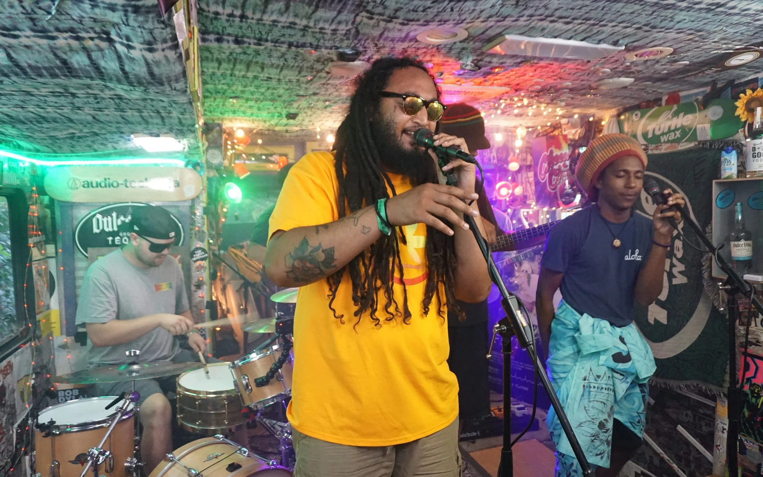 Dread Kennedy singing into a microphone at a lively indoor music venue with colorful lights. Nearby, a drummer wearing a black beanie and glasses is playing drums, and a young man in a striped beanie and blue shirt is holding a microphone.