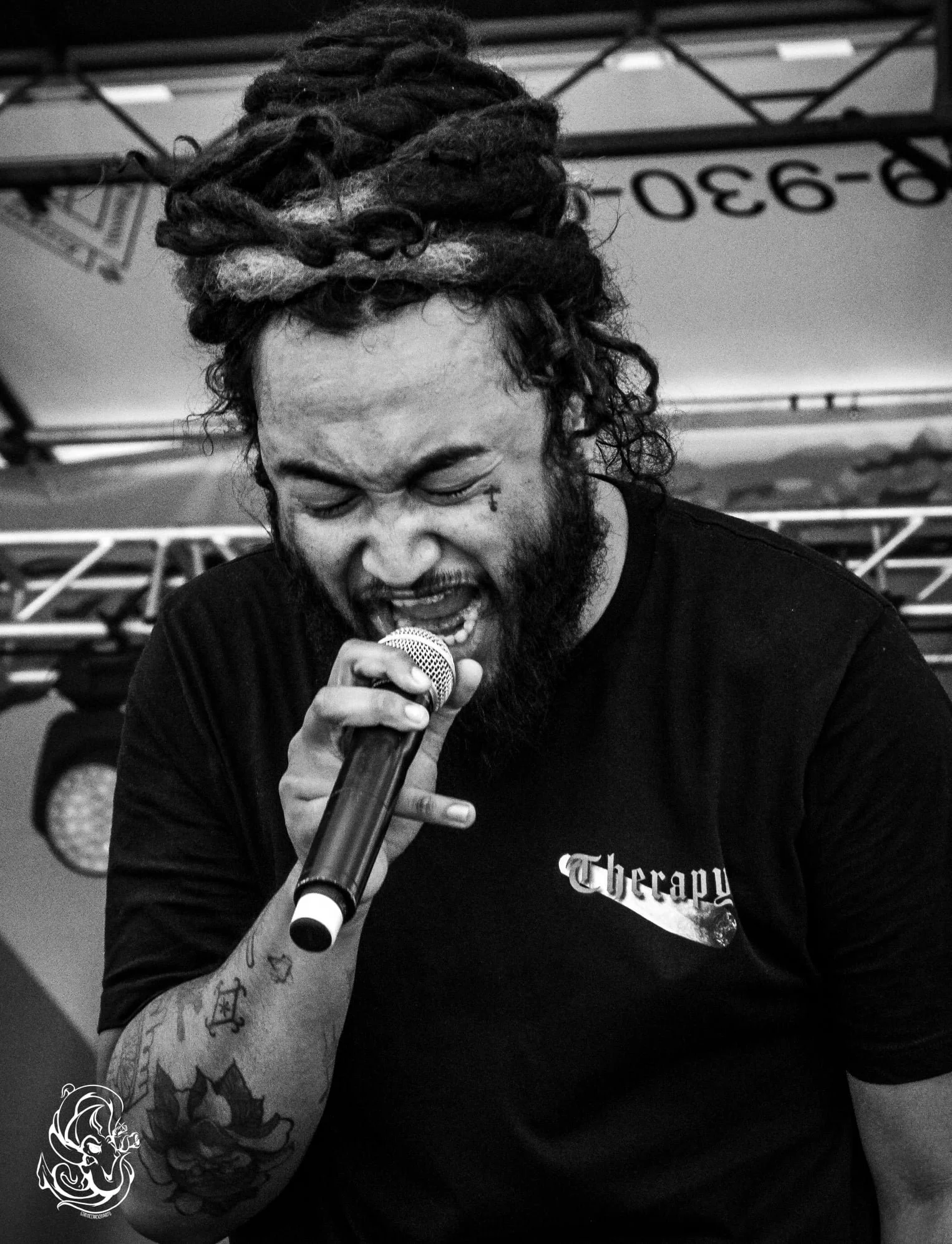 A man with dreadlocks and tattoos on his arm is passionately singing into a microphone at a live music event.