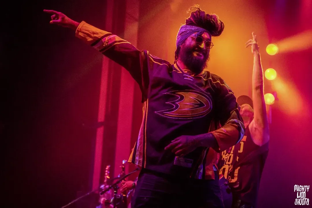 A man with dreadlocks, wearing sunglasses, a colorful sports jersey, and a head wrap, is dancing on stage with colorful lighting.