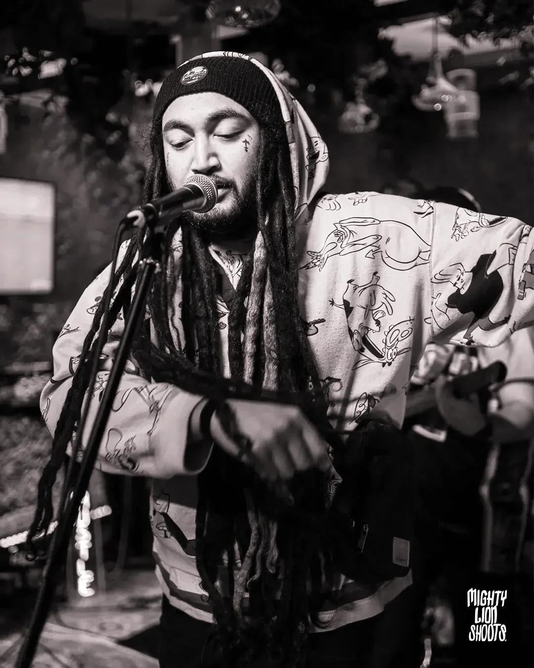 Dread Kennedy wearing a beanie and a hoodie with cartoon characters, singing into a microphone, performing on stage with a guitar in a dimly lit setting.