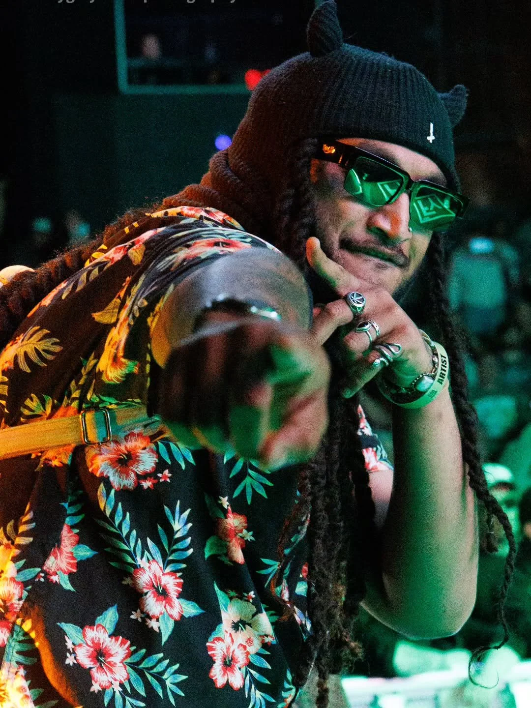 Dread Kennedy in sunglasses, a black beanie with snout and ear details, tropical floral shirt, ring, and bracelets, pointing directly at the camera in a dark setting with colorful lighting.