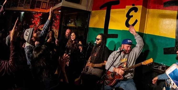 People enjoying live music performance in an indoor venue with a rainbow-colored flag in the background.