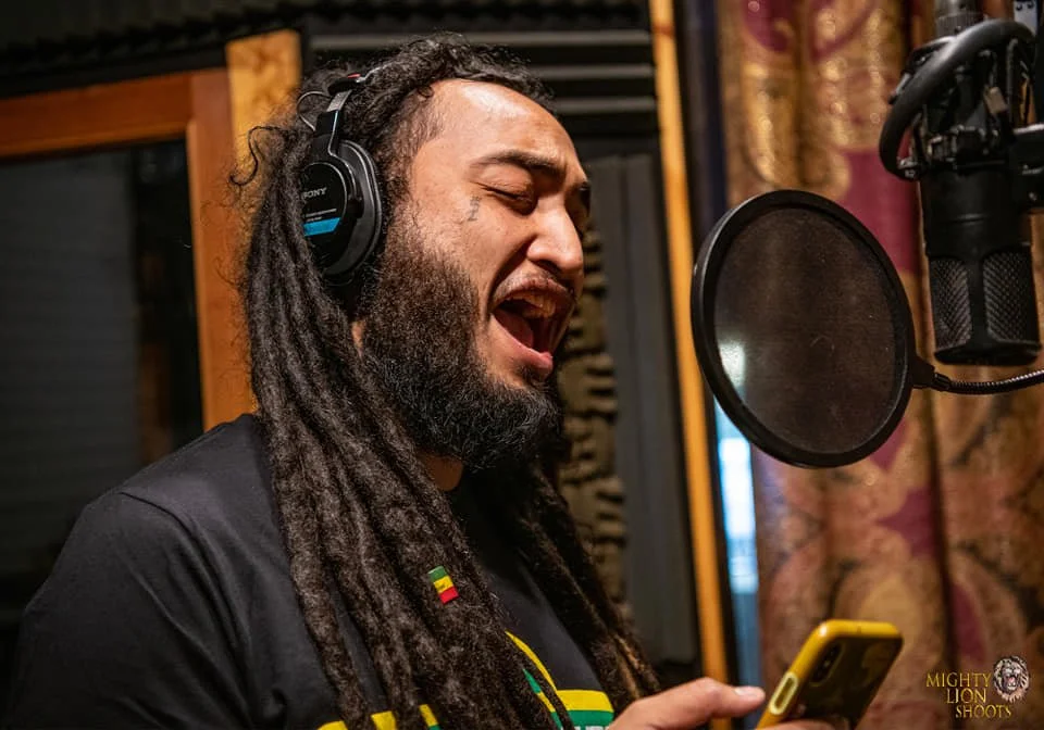 Man with long dreadlocks singing into a microphone in a recording studio, wearing headphones and holding a smartphone.