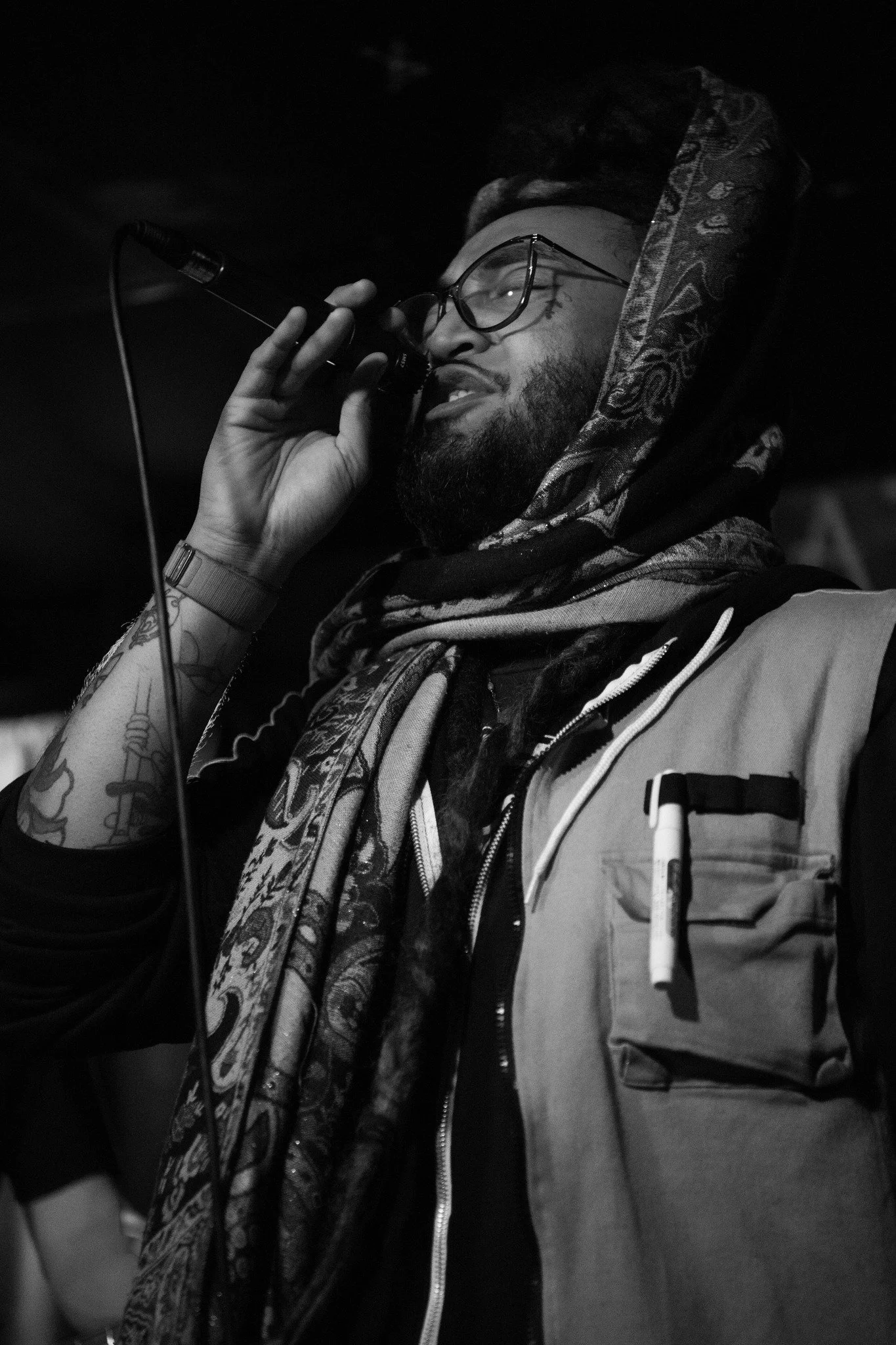A man with glasses drinking from a microphone, wearing a scarf and a jacket with a pen in the pocket, tattoos on his arm, in a dark setting.
