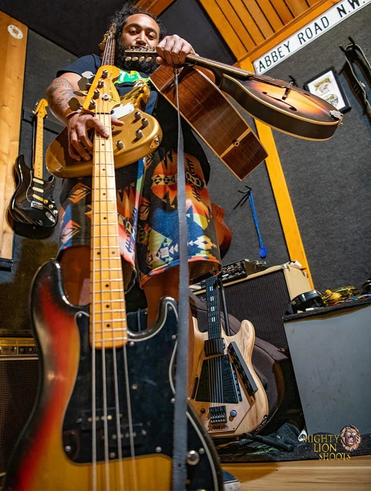 Dread Kennedy in a colorful shirt holding a guitar above several other guitars in a music studio.