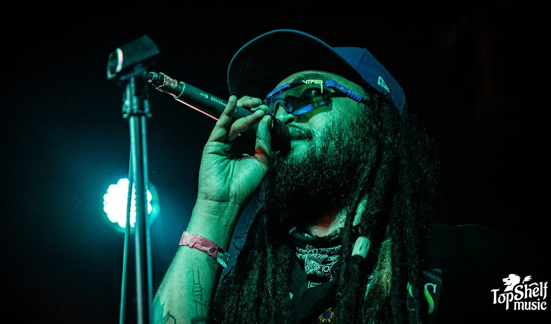A male performer with long dreadlocks, wearing glasses, a baseball cap, and a bandana around his neck, singing into a microphone on stage with blue and green stage lighting.