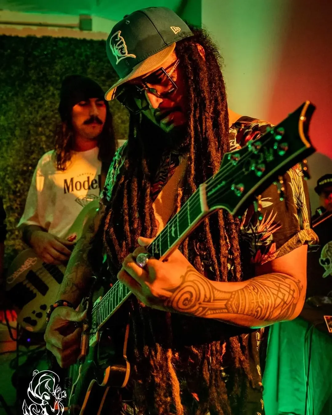 Dread Kennedy performing with guitars under colorful stage lighting, one wearing a cap and sunglasses, tattooed arm visible.