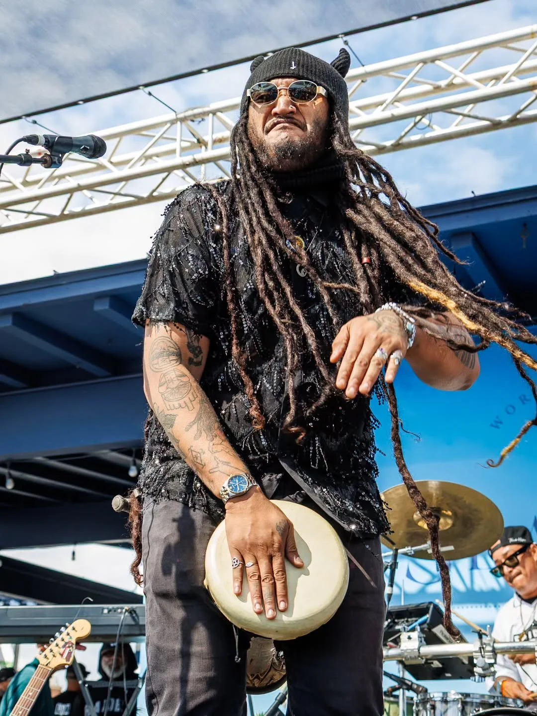 Dread Kennedy wearing sunglasses and a beanie playing hand drums on stage at an outdoor concert.