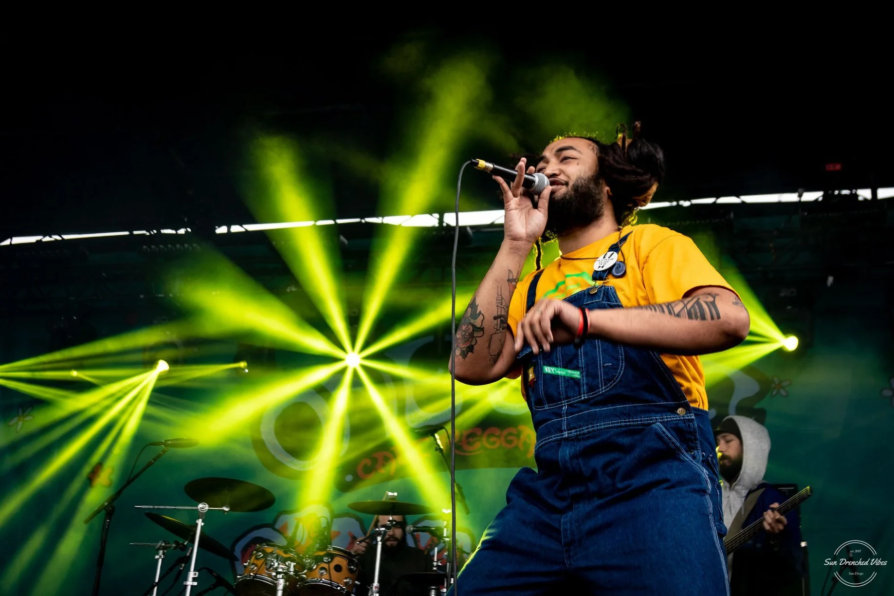 Dread Kennedy singing into a microphone on stage with bright yellow stage lights in the background, wearing a yellow shirt and denim overalls, with tattoos on arms.