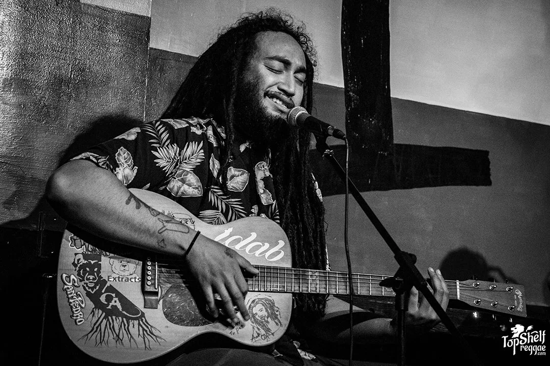 Dread Kennedy wearing a floral shirt, is singing into a microphone while playing an acoustic guitar with various stickers and drawings on it. He appears to be performing passionately on stage.