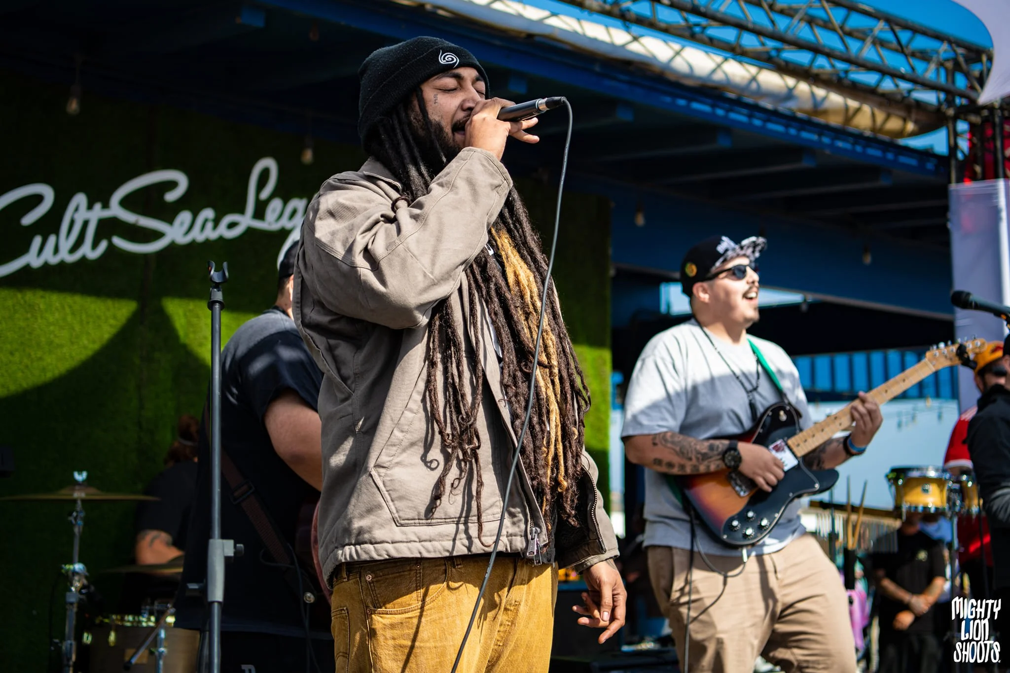 Dread Kennedy singing into a microphone on an outdoor stage. Other band members are visible in the background, one playing an electric guitar and wearing sunglasses. The stage is set against a green backdrop with a sign that reads 'Cult SeaLegs'.