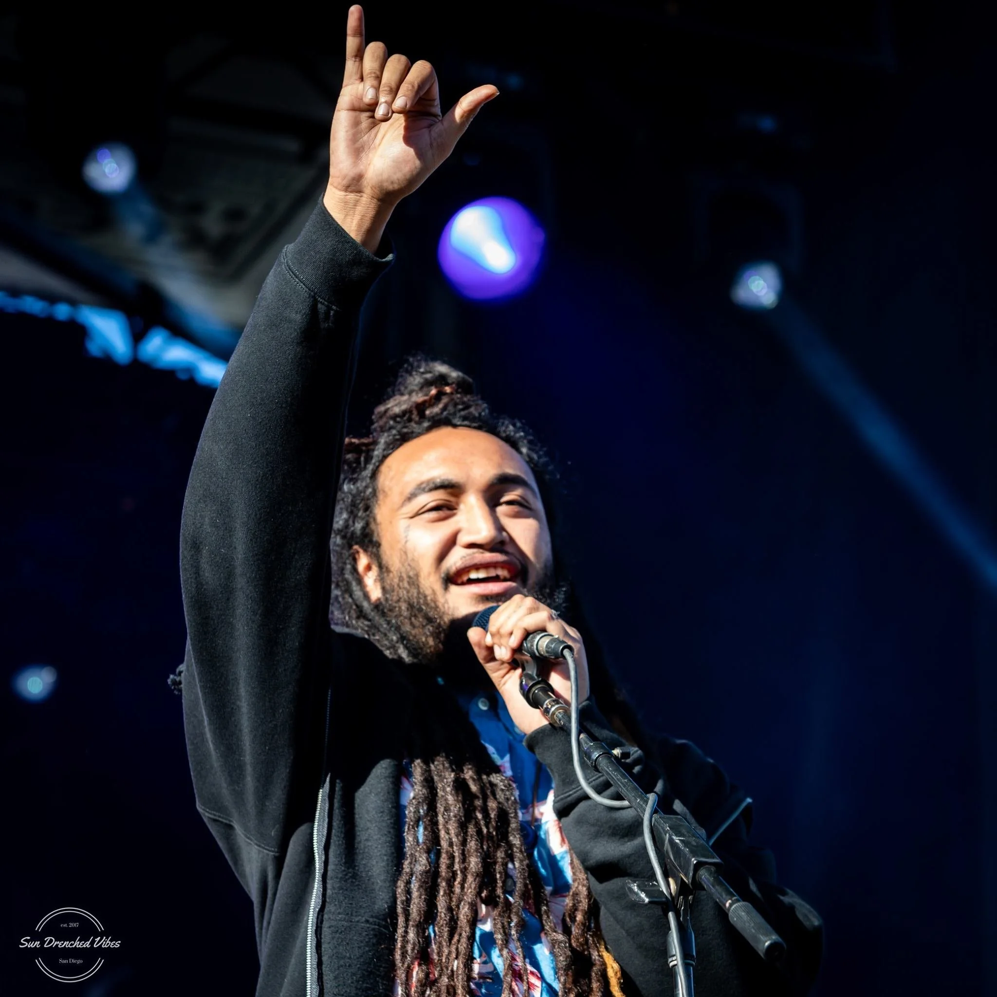 Dread Kennedy smiling and raising their hand with a finger pointing up on stage, holding a microphone, with dark background and stage lights.