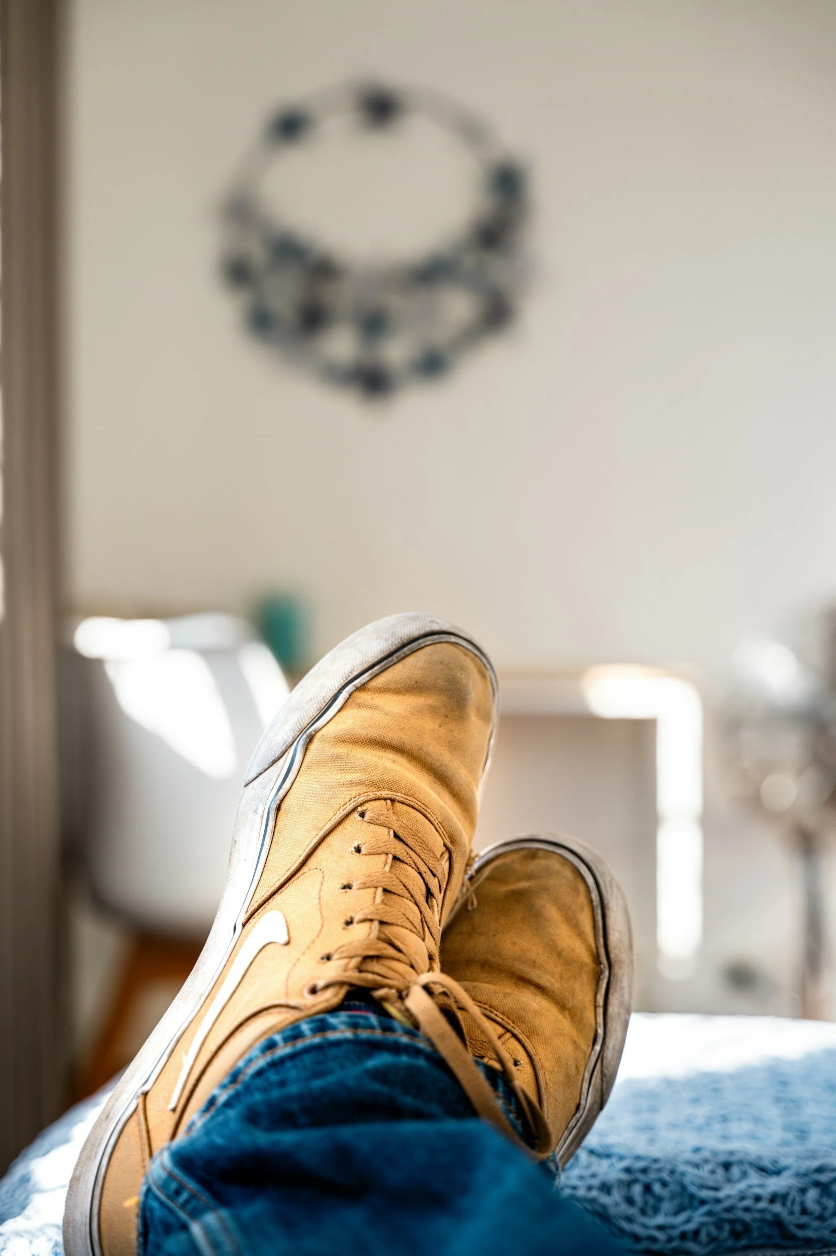 Person wearing tan sneakers and blue jeans relaxing with legs extended in a room with sunlight, with blurred hanging décor and a window in the background.