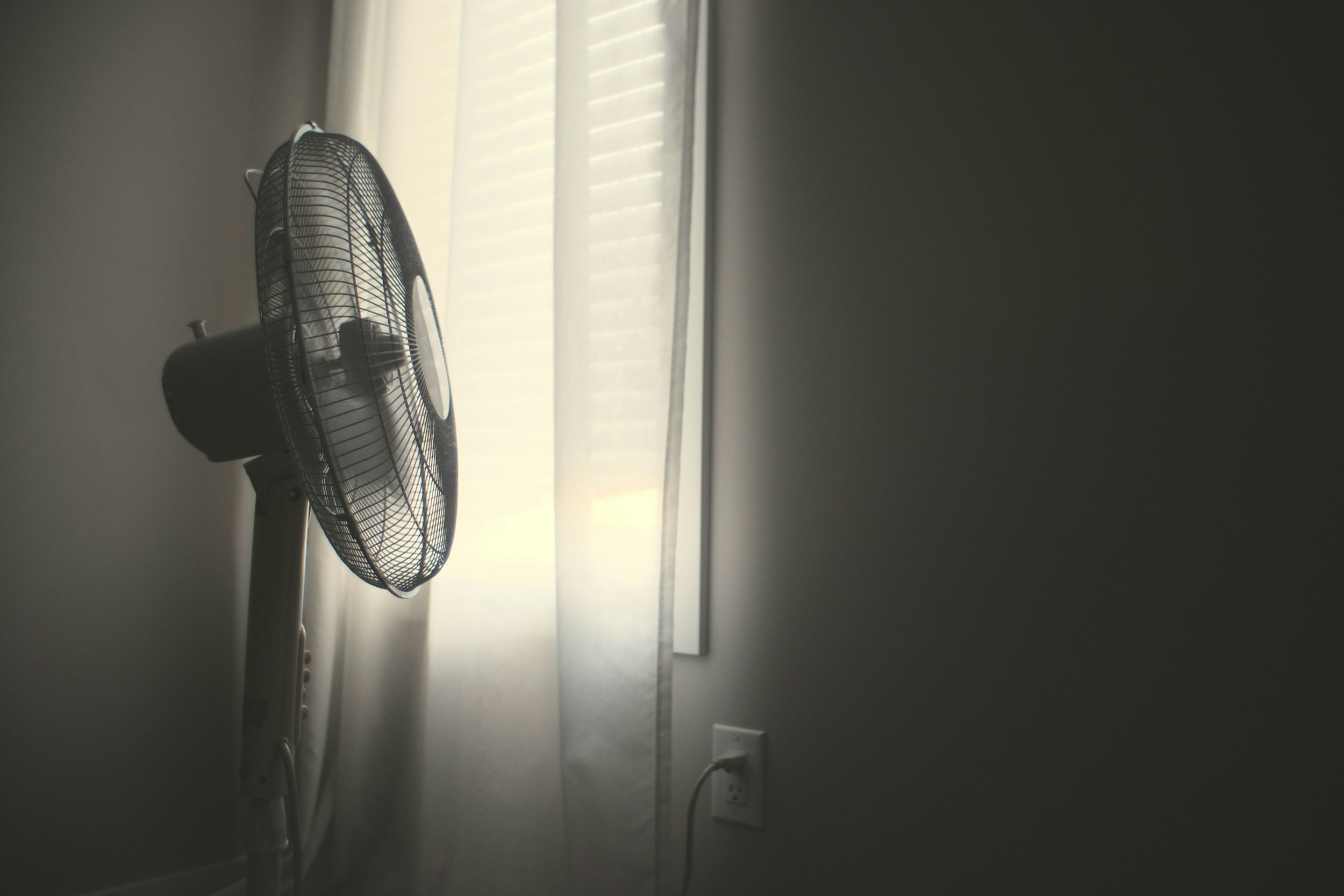 A black oscillating fan positioned in front of a window with sheer curtains, in a room with plain walls and a visible electrical outlet.