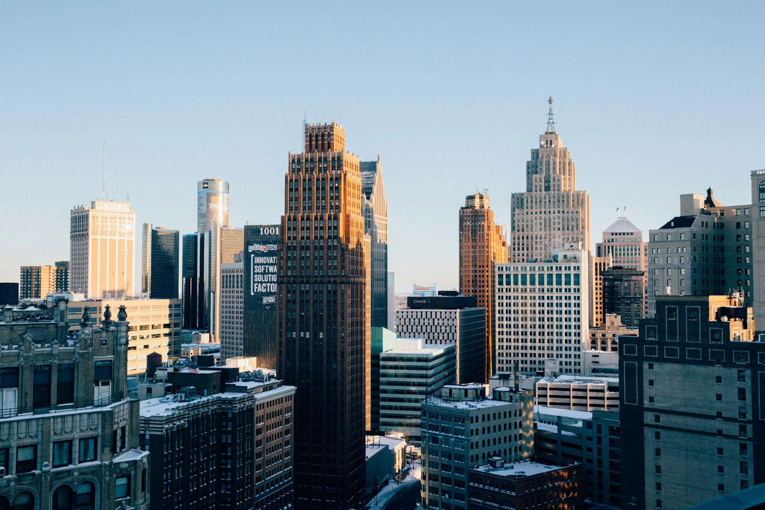 Skyline of a city with tall buildings, some with art deco architecture, under a clear blue sky, with snow on rooftops.