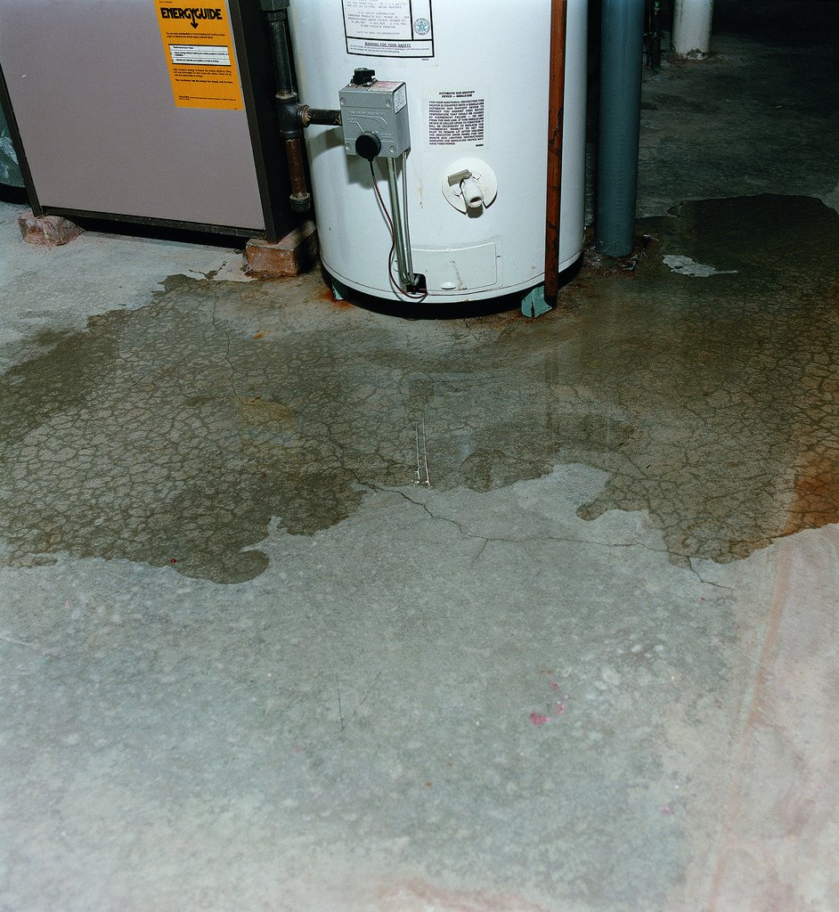 A water heater and a furnace or boiler in a basement or utility room, with wet stains on the concrete floor.