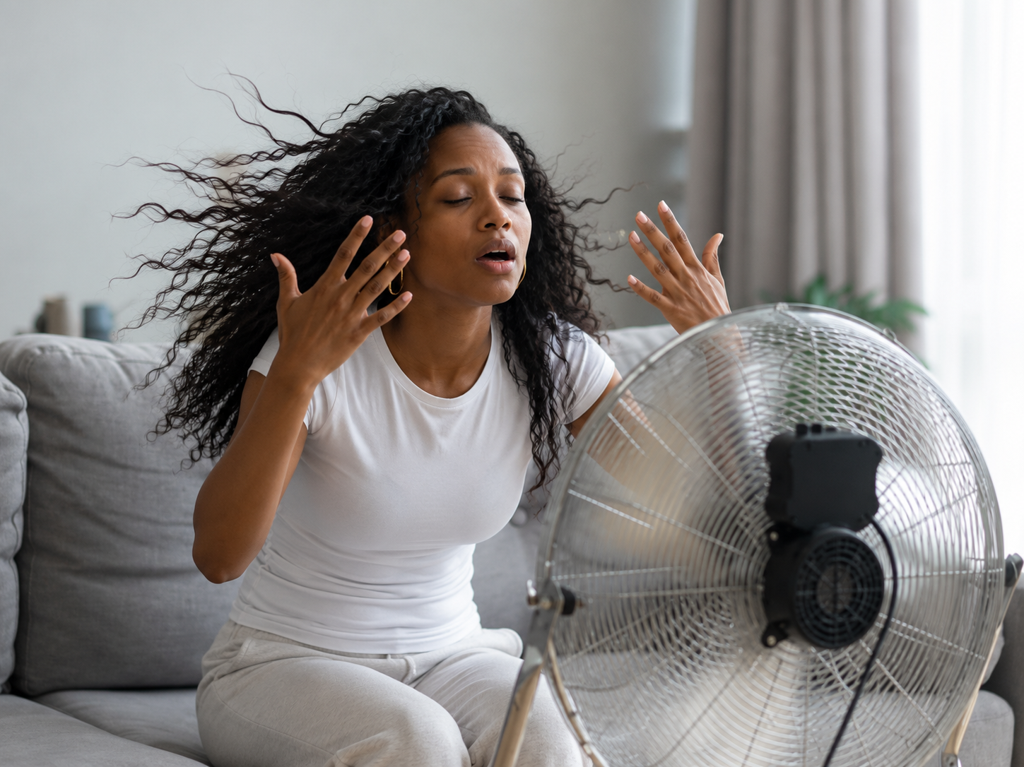 A woman with curly hair, wearing a white t-shirt, sitting on a couch and sweating with her eyes closed while blowing on a fan in a living room.