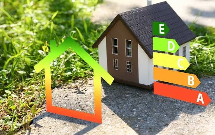 A small model house with an energy efficiency rating chart showing ratings from A to E, placed outdoors on the ground near green grass.