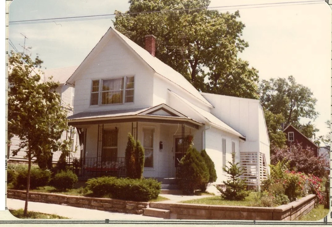 A white two-story house with a gable roof, front porch, and front yard with trees and shrubs, in a suburban neighborhood.